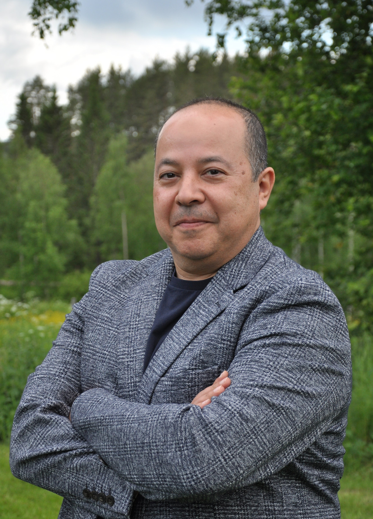 A man with a grey suit. He is outside, green woods in the background.